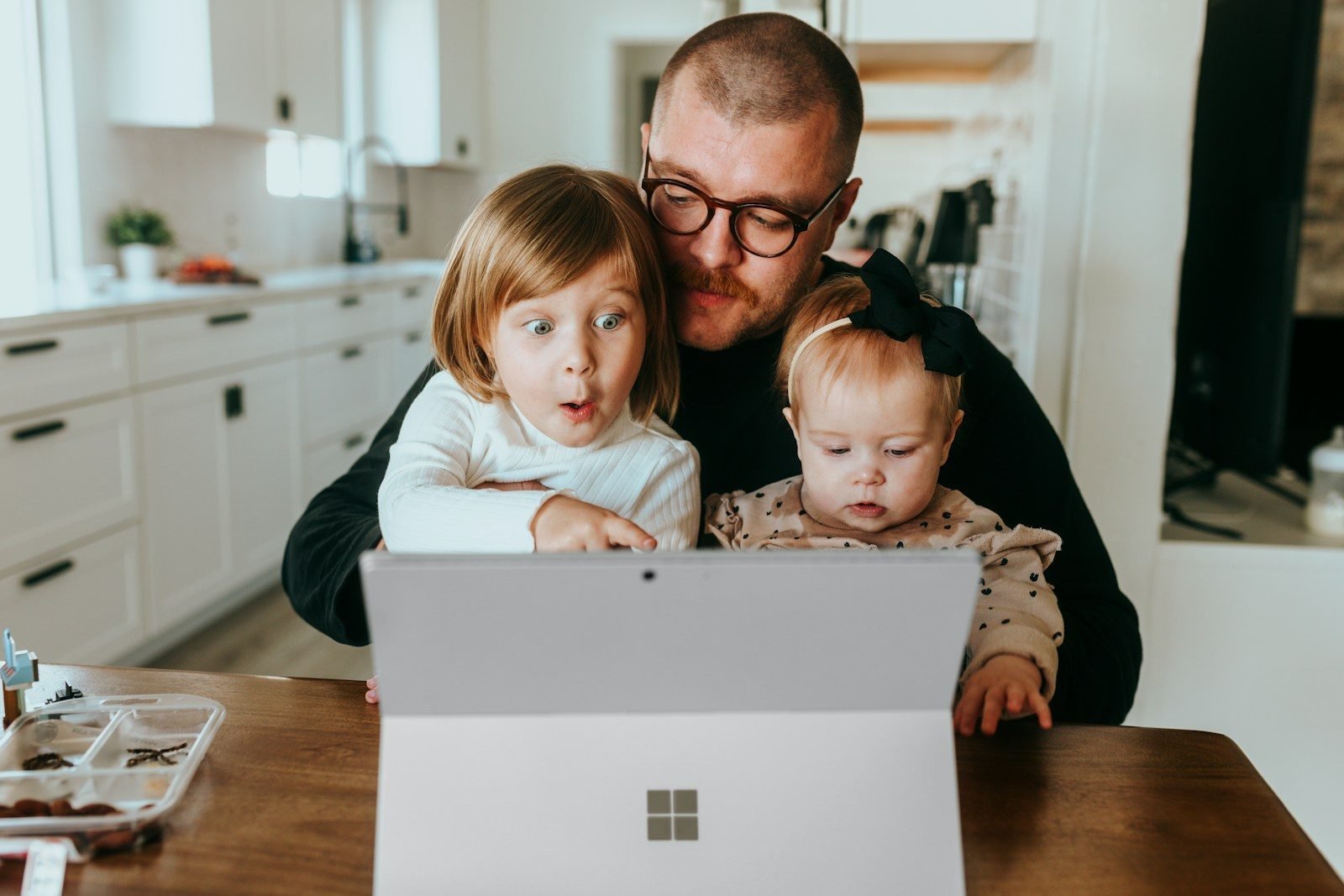 A father at the kitchen table with his toddler and his little girl, all three looking at his laptop
